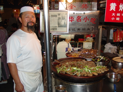 Kok bij een eetkraam op de Zijderoute, met grote pan streetfood en Chinese uithangborden.