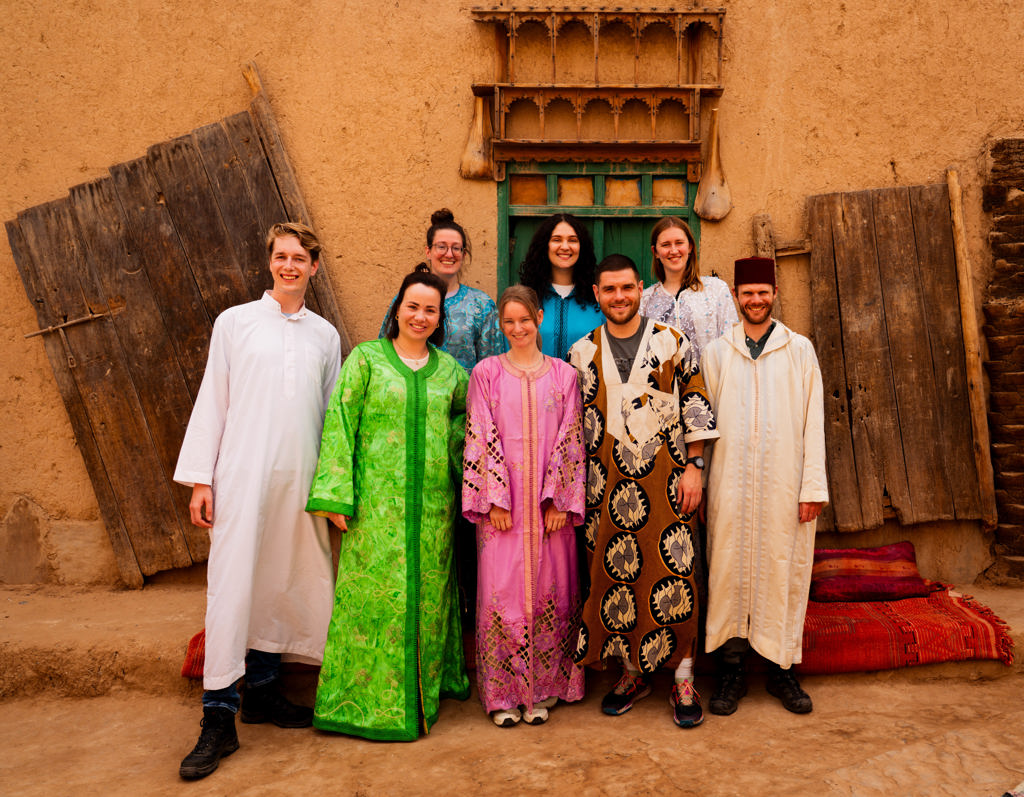 Groep 22-35ers poseert in traditionele Marokkaanse kleding bij een lemen huis tijdens een rondreis door Marokko.