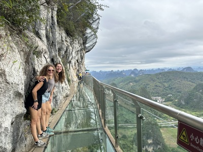 Twee reizigers op een glazen skywalk langs een rotswand met uitzicht over groene bergen en dal.